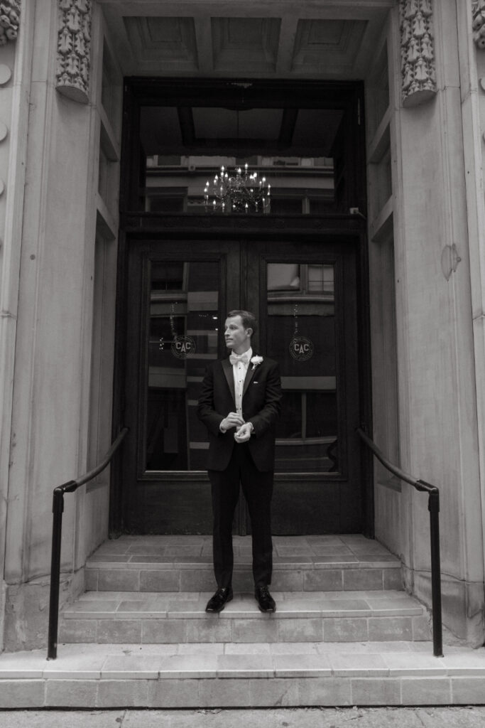 Groom stands on steps outside Cincinnati Athletic Club before cozy Christmas wedding ceremony.