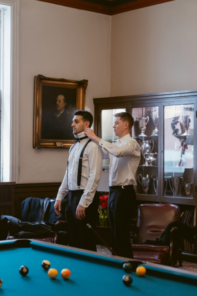 Groomsmen adjusting bowtie in vintage billiards room before cozy Christmas wedding celebration.