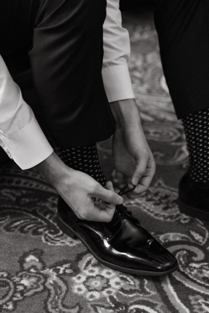 Close-up of groom tying black dress shoes, wearing polka dot socks, on patterned carpet before cozy Christmas wedding.