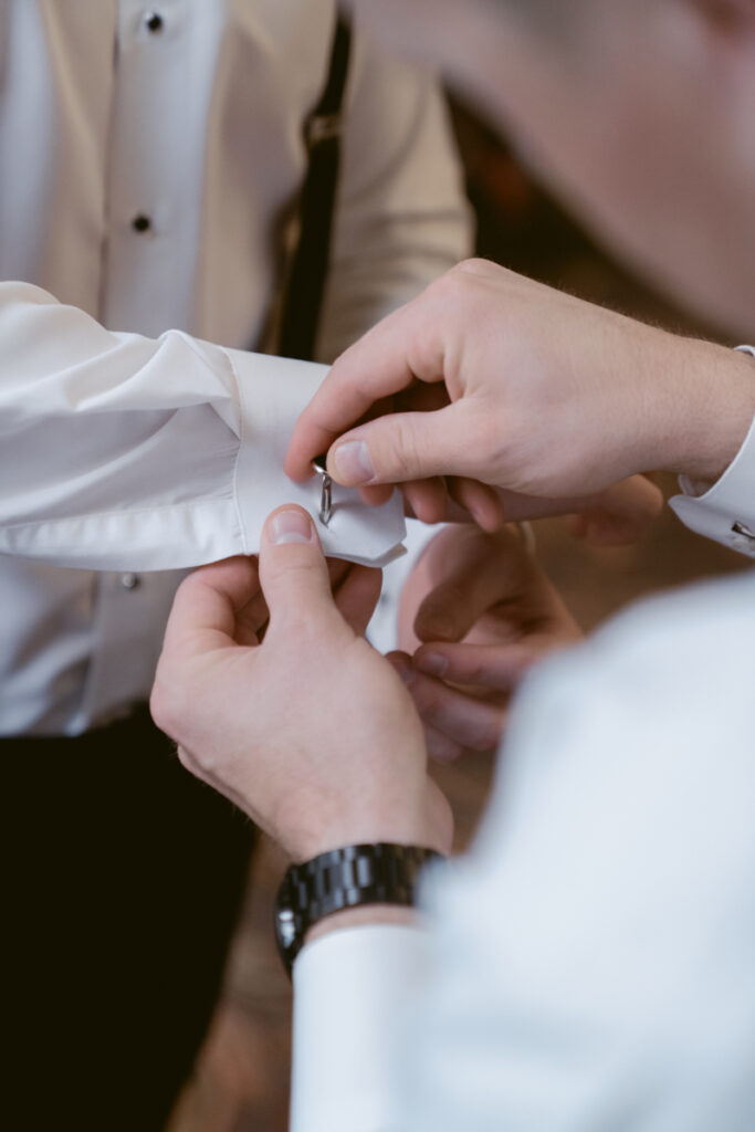 Groomsman fastening cufflink on groom's white dress shirt before cozy Christmas wedding.