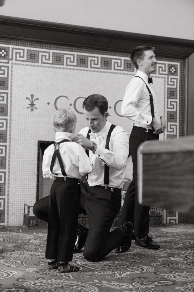 Groom kneeling to help ring bearer with his bow tie while getting ready for a cozy Christmas wedding, with groomsmen in the background.