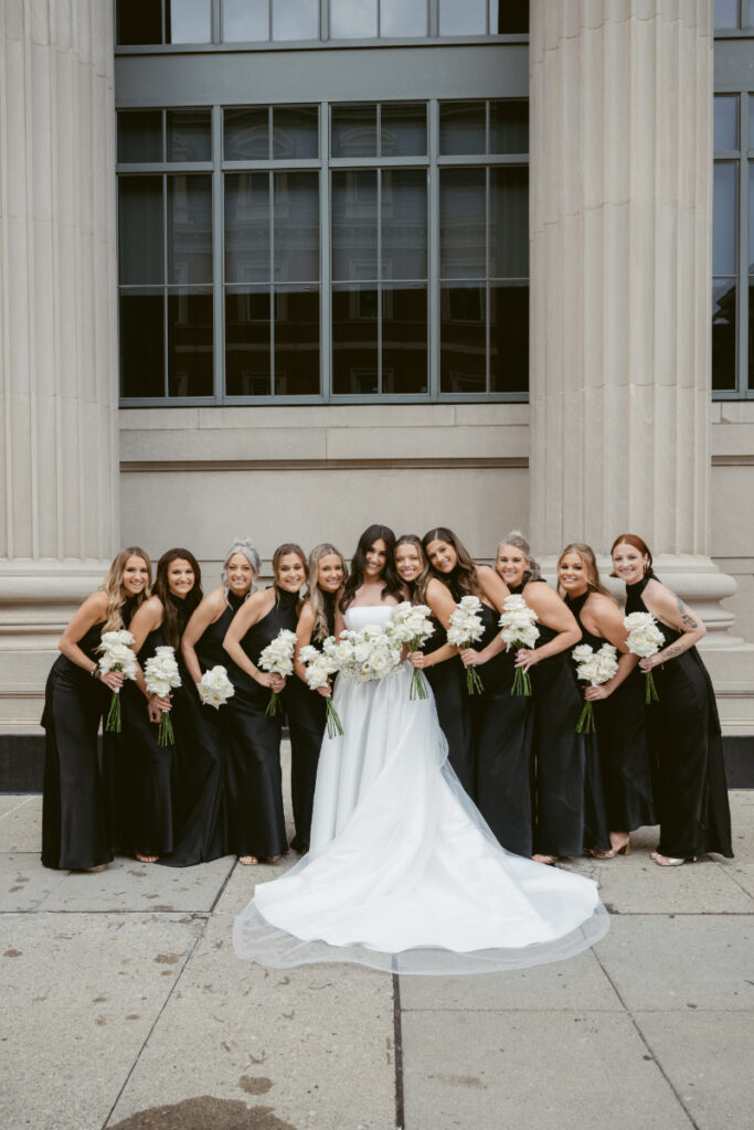 Bride surrounded by bridesmaids in black dresses, holding white bouquets outside a grand city building.