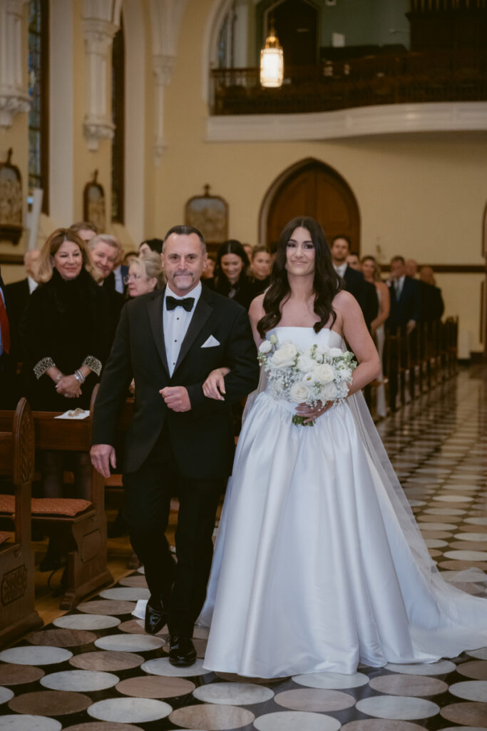 bride walking down the aisle with her dad, guests are standing and watching 