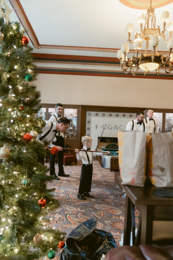 Ring bearer plays pool with groomsmen in a decorated lounge featuring a Christmas tree, ahead of a cozy holiday wedding.