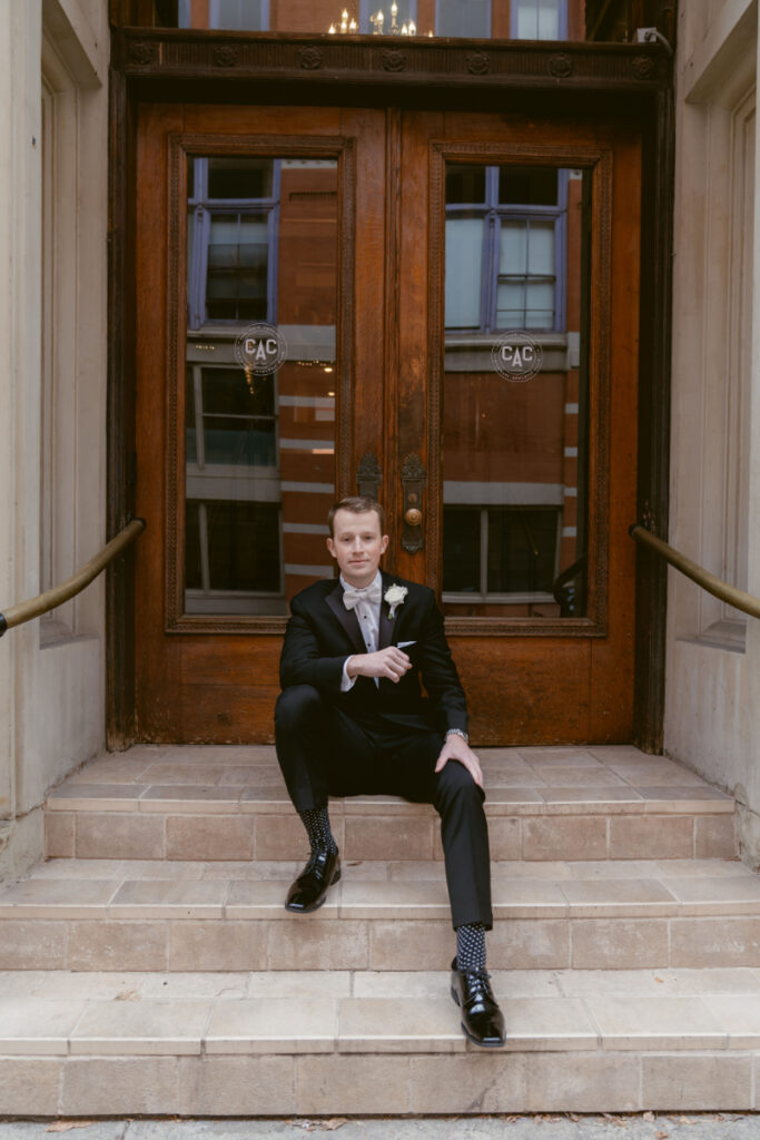 Groom sits on the steps outside the Cincinnati Athletic Club, dressed in a black tuxedo with bow tie, looking relaxed before the ceremony.