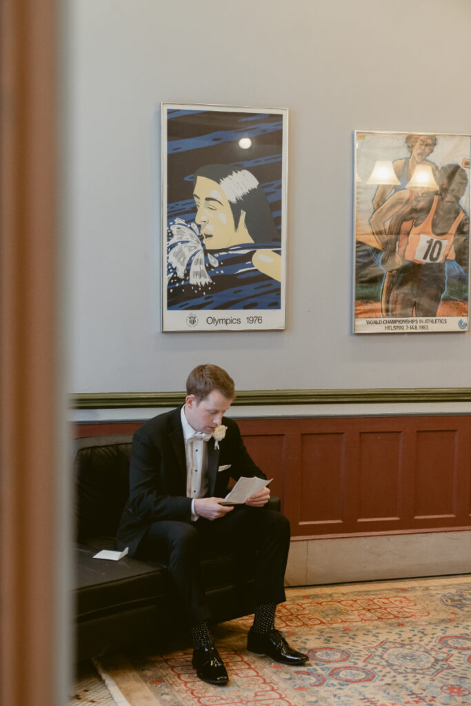 Groom in tuxedo sits on a black leather bench reading a letter, with vintage Olympic posters hanging on the wall behind him.