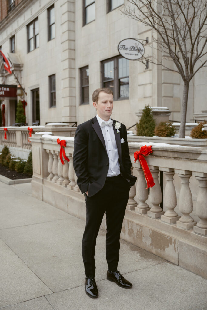 Groom in a black tuxedo standing on a city sidewalk in front of The Phelps hotel, with festive red bows decorating the stone railing.