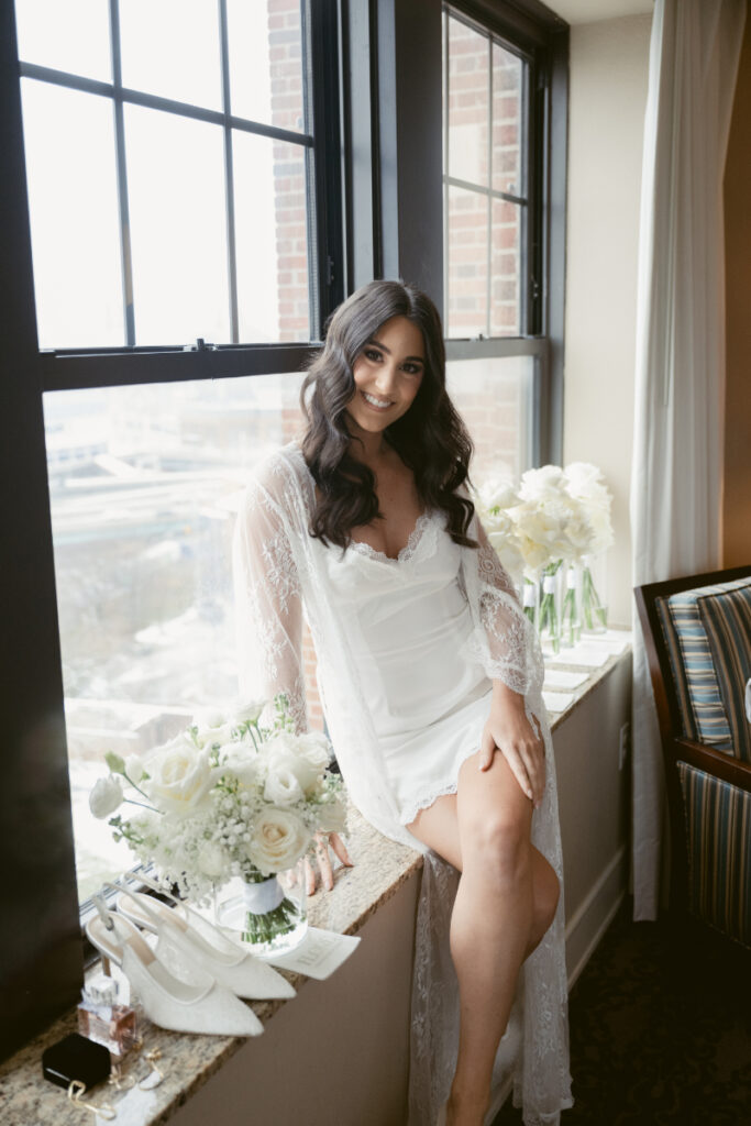 Bride sitting by window in robe on cozy Christmas wedding morning, with bouquet and bridal details.