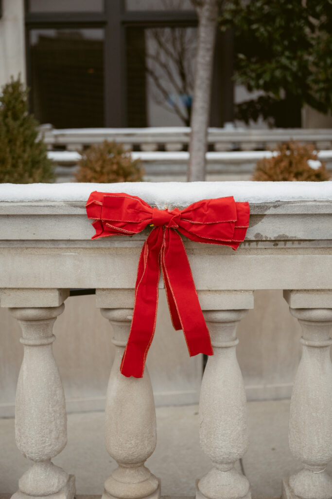 A bright red velvet bow tied to a snowy stone railing adds festive cheer.