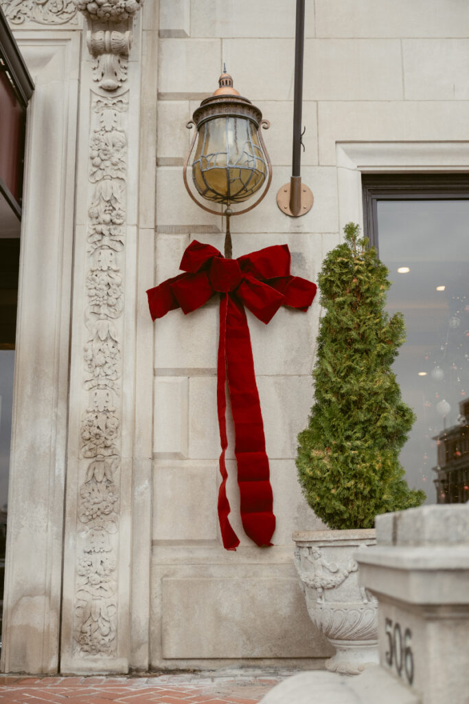 Large red velvet bow hanging under a vintage-style lantern on a stone building exterior, with a small evergreen tree in a white planter nearby.