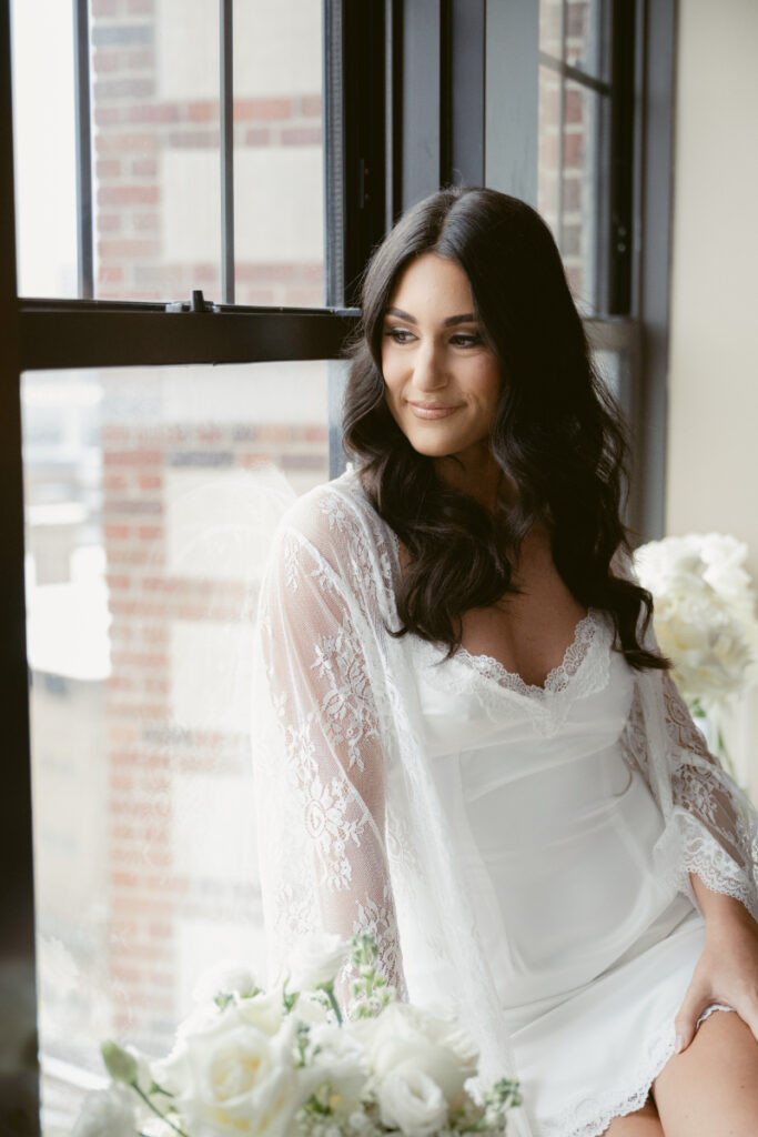 Bride in lace robe sitting by window on cozy Christmas wedding morning, surrounded by white roses.