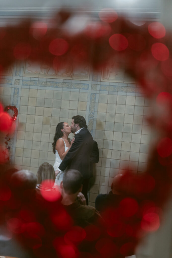 Couple kissing framed by red heart decor at Valentine’s Day wedding.