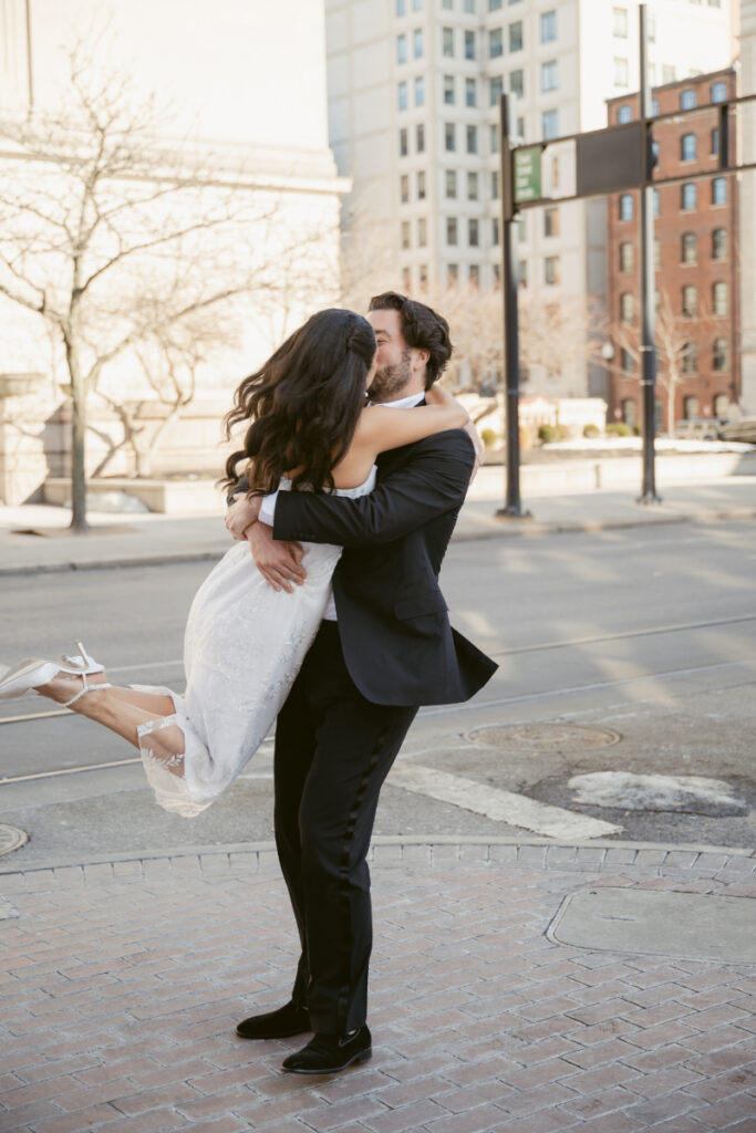 Groom lifting bride in street during joyful Valentine’s Day wedding portraits.