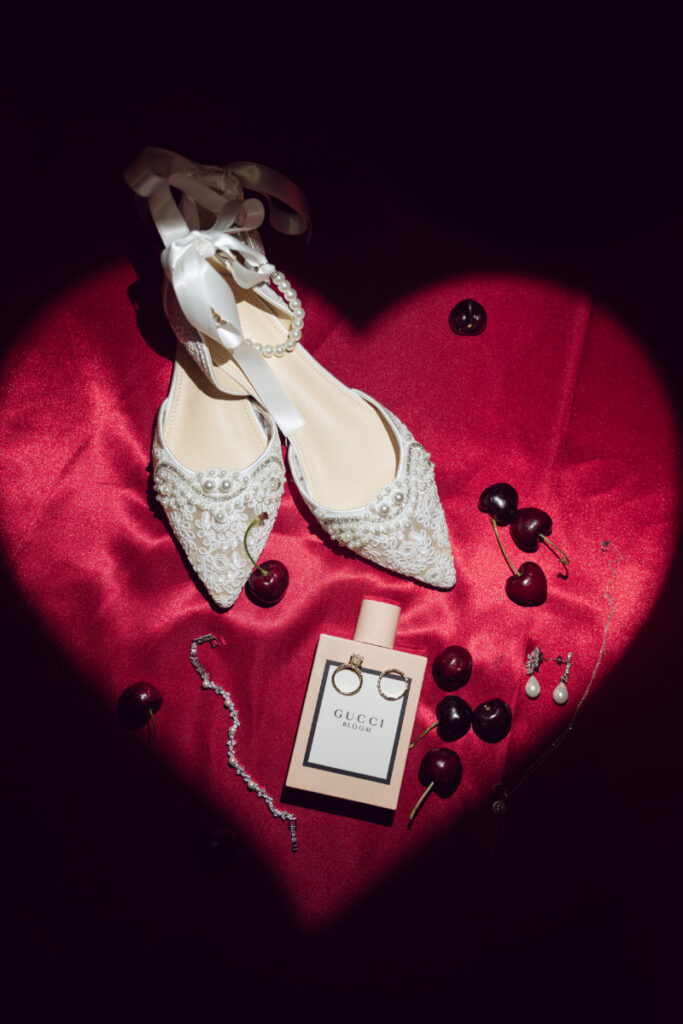 Flat lay of bridal shoes, cherries, and perfume on red velvet fabric.