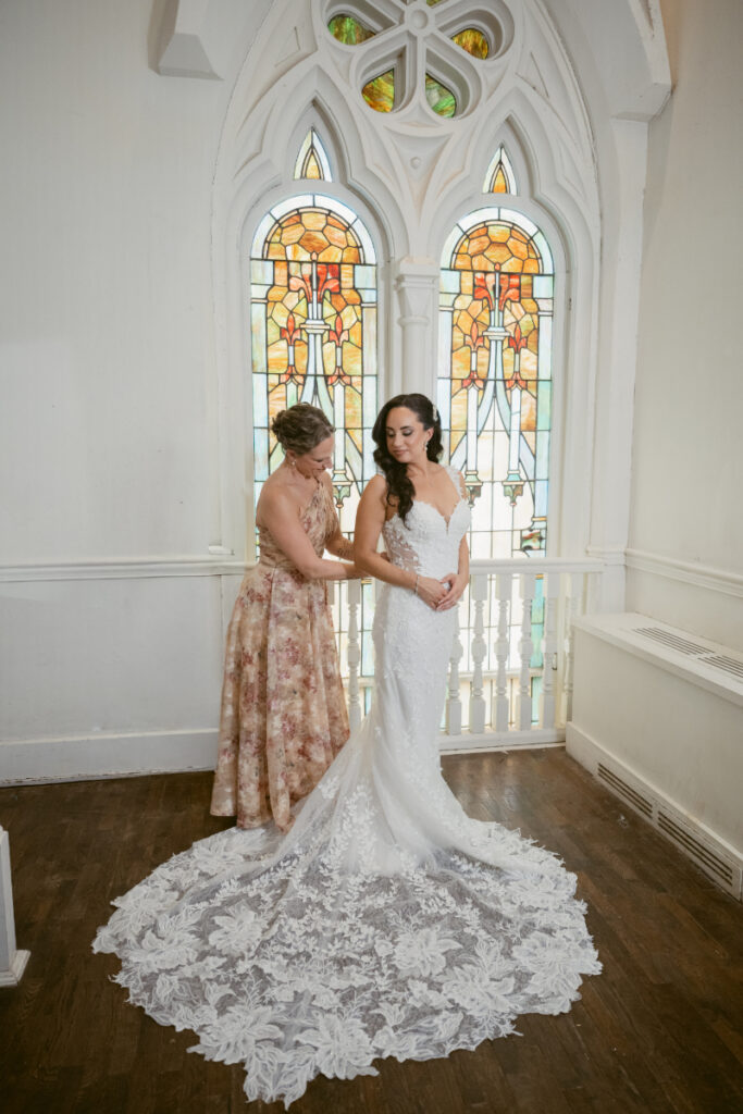 Bride standing with mother near stained glass window before ceremony.