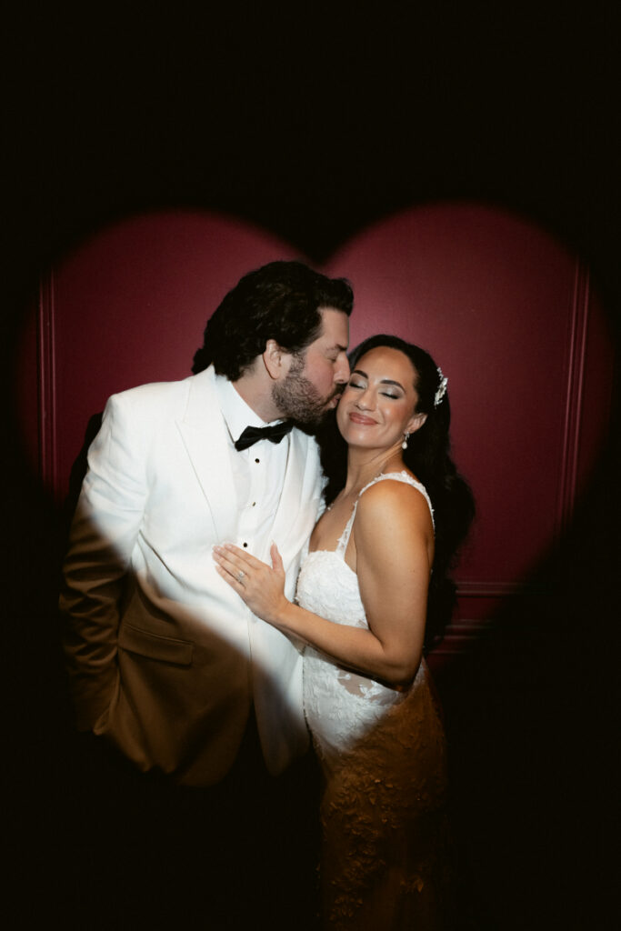 Bride smiling as groom kisses her cheek against red heart backdrop.