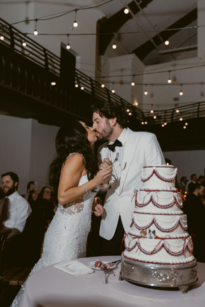 Bride and groom kissing while cutting red and white tiered wedding cake.
