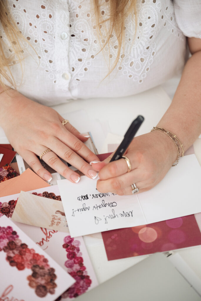 Close-up of hands writing on a card surrounded by colorful stationery and floral paper designs.