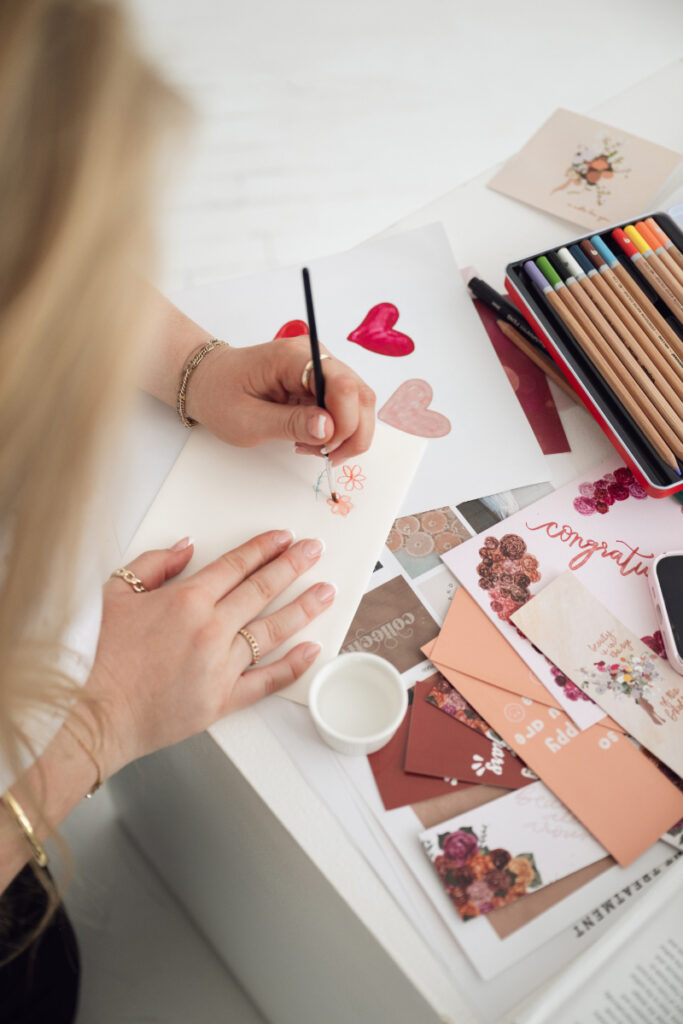 Overhead view of a woman sketching heart designs on stationery, surrounded by colorful cards and art supplies.