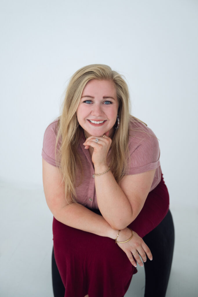 Smiling woman leaning forward with hand under chin, wearing a pink top and red skirt against a white background.