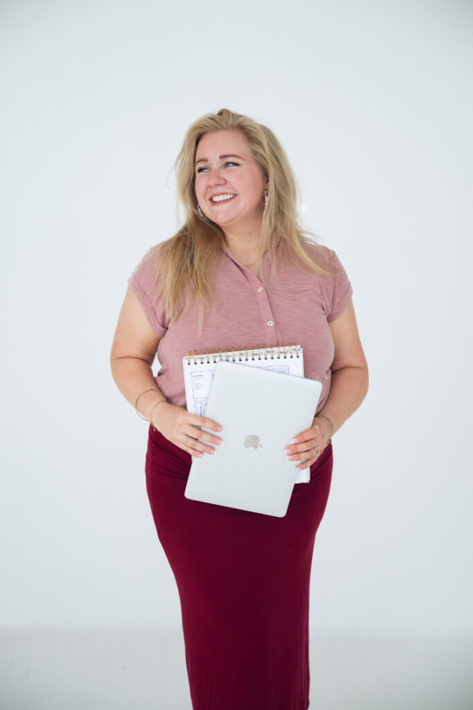 Smiling woman in pink top holding laptop and planner against a clean white background.