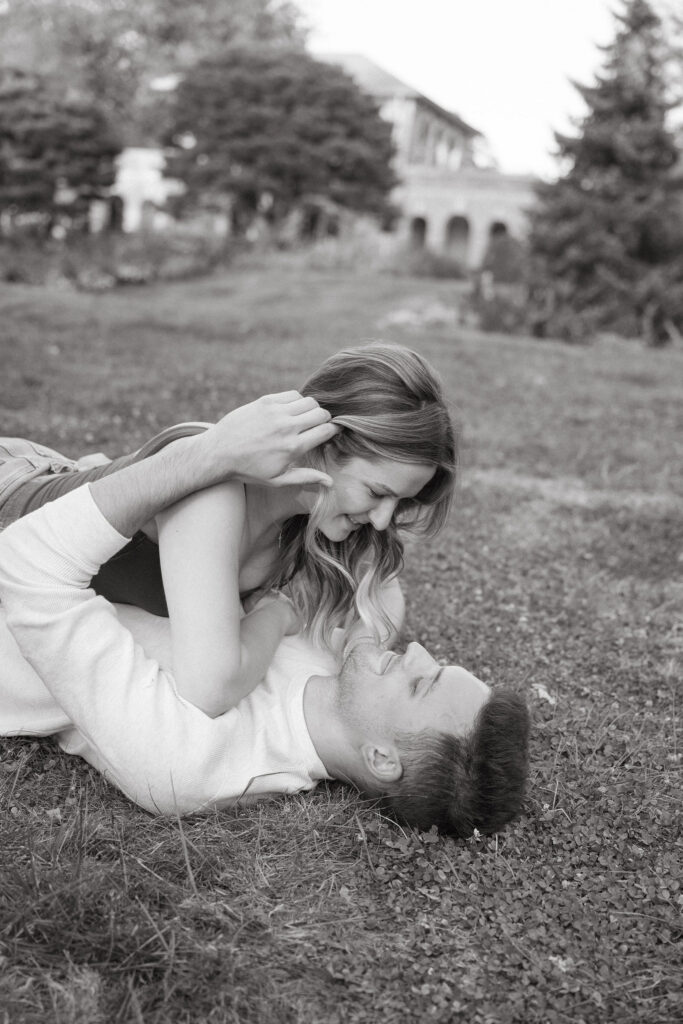 A couple lies on the grass laughing together, faces close and playful. The candid moment captures joy and comfort, making for relaxed, authentic natural engagement photos with a lighthearted feel.