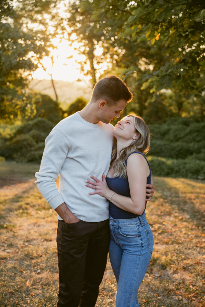 A couple stands close together in golden hour light, gazing into each other’s eyes. Warm tones and soft shadows highlight their connection, creating timeless natural engagement photos in a peaceful outdoor setting.