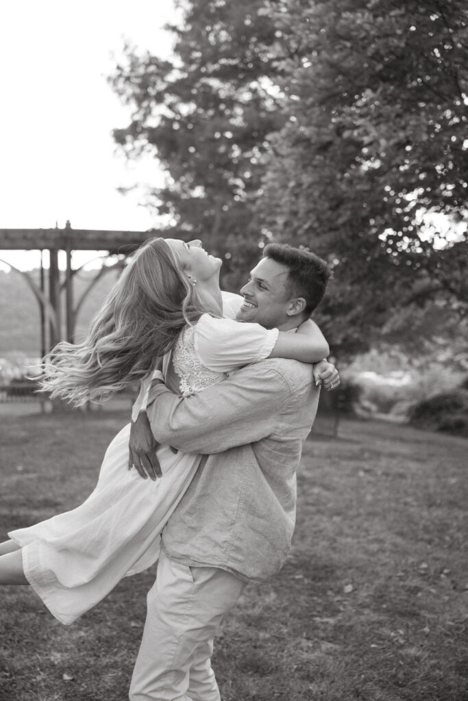 In a black-and-white image, a man lifts his partner as she laughs, hair flowing mid-motion. A bridge and trees blur behind them, capturing carefree energy and connection—classic natural engagement photos with movement and emotion.
