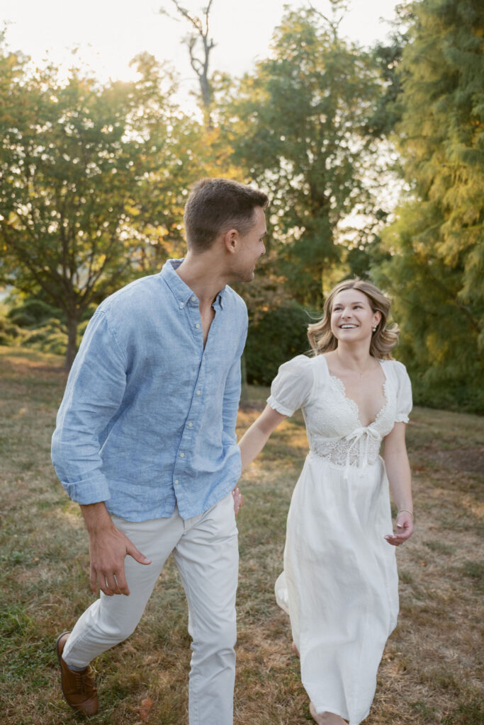 A couple walks hand in hand through a sunlit field, smiling at each other. Soft greenery and warm tones surround them, creating natural engagement photos that feel effortless, romantic, and full of motion.
