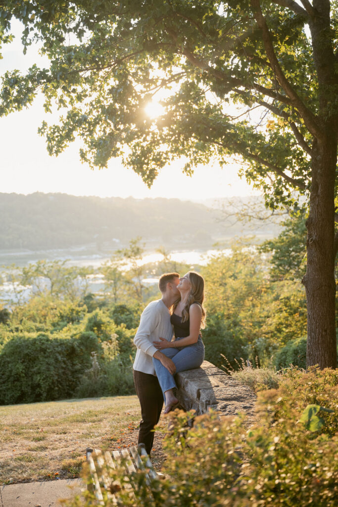 A couple sits and stands along a stone ledge under a glowing sunset, framed by leafy branches. Golden light filters through, creating a warm, dreamy atmosphere—romantic natural engagement photos overlooking a scenic landscape.