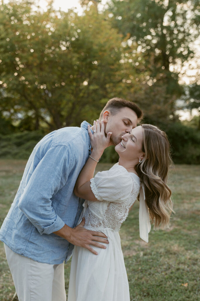 A couple shares a playful, affectionate moment outdoors as he leans in to kiss her cheek. She smiles with eyes closed, hand on his neck, surrounded by soft greenery and warm light—romantic natural engagement photos with a candid, joyful feel.