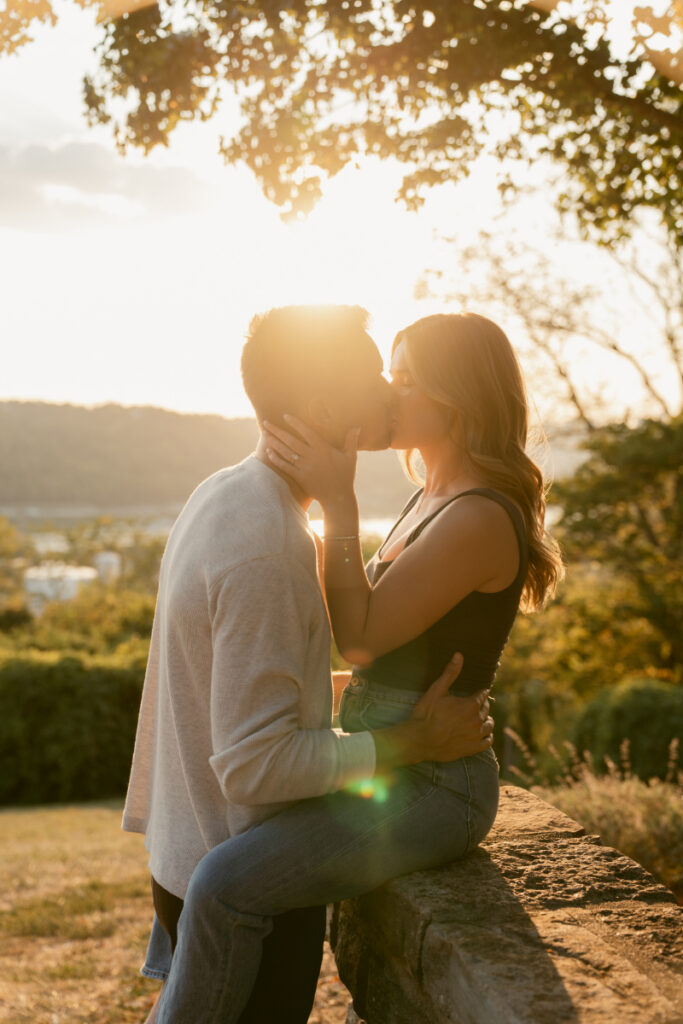 Couple kissing in golden hour light with soft sun flare and scenic overlook in background