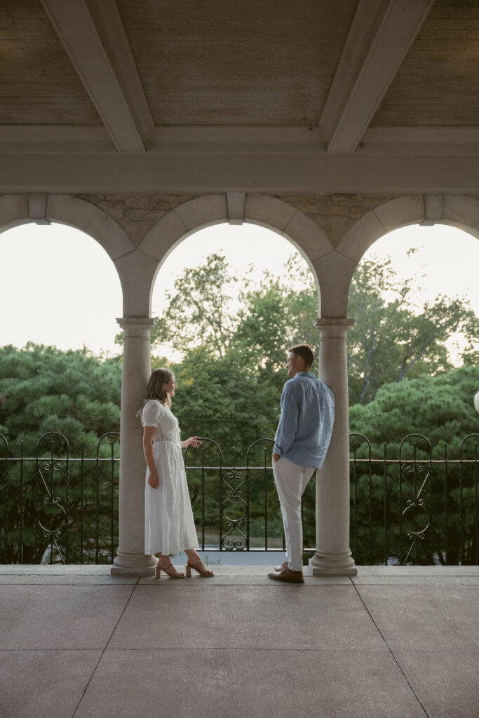 A couple stands facing each other beneath stone arches on a terrace, framed by columns and lush trees beyond. The scene feels calm and intimate, with soft evening light creating timeless, natural engagement photos in an elegant architectural setting.