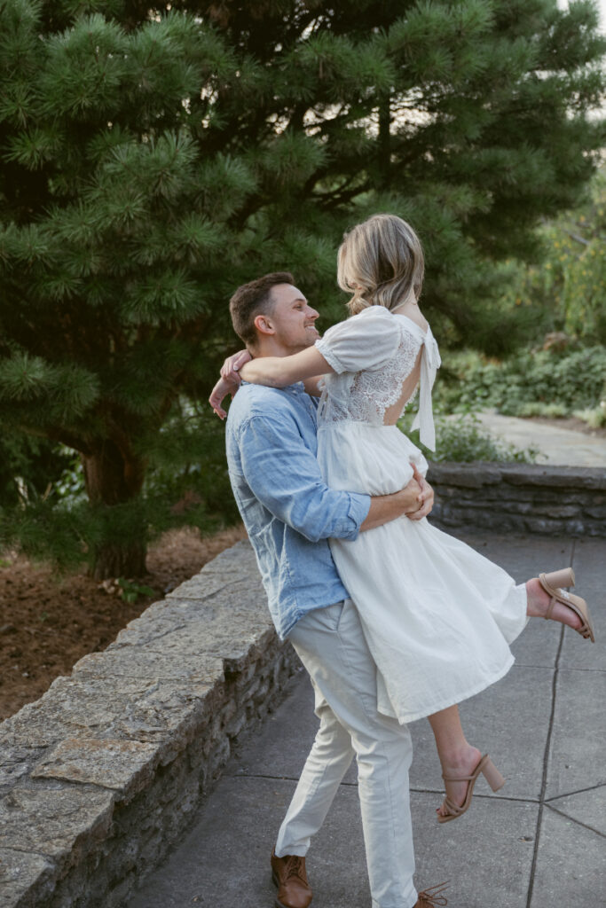 A man lifts his partner near a stone wall as she wraps her arms around him, both smiling. Surrounded by greenery, the moment feels playful and intimate—lighthearted natural engagement photos with a relaxed, candid vibe.