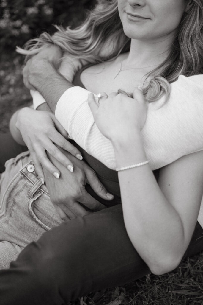 A close-up black-and-white image of an embrace focuses on hands, jewelry, and connection. The tight framing creates an intimate mood—refined natural engagement photos emphasizing emotion and detail.