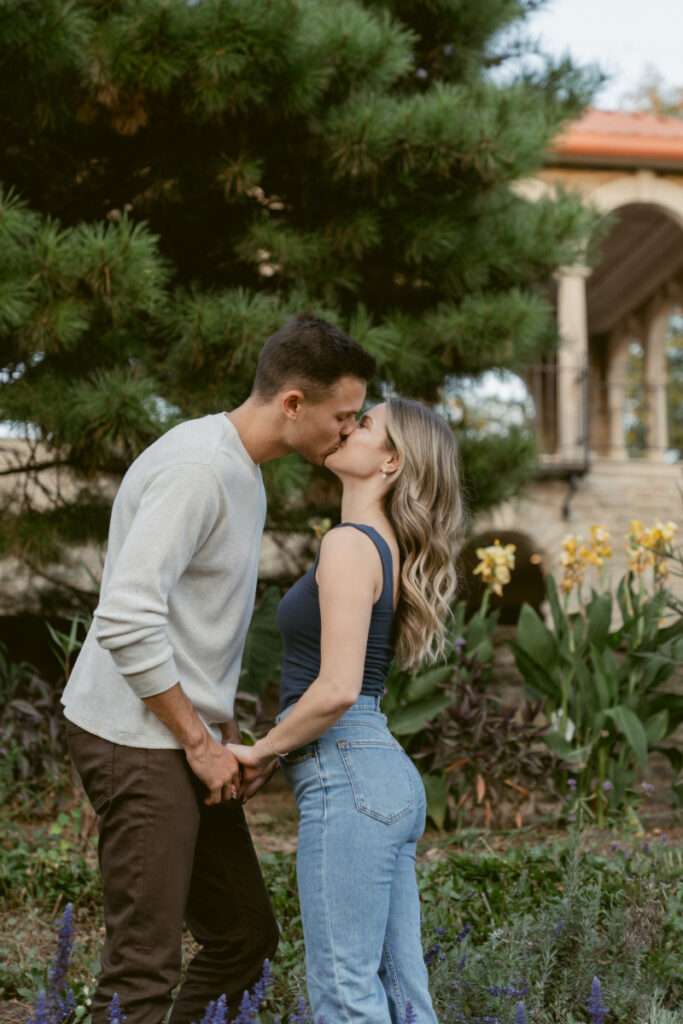 The couple shares a kiss while standing in a lush garden with flowers and greenery. The soft background and gentle pose create a tender, intimate moment—beautiful natural engagement photos with a garden setting.