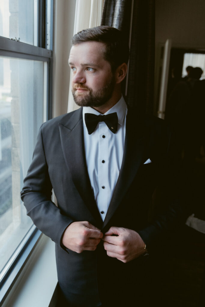Groom adjusting his jacket by a window during getting ready at a Hilton Netherland Plaza wedding.