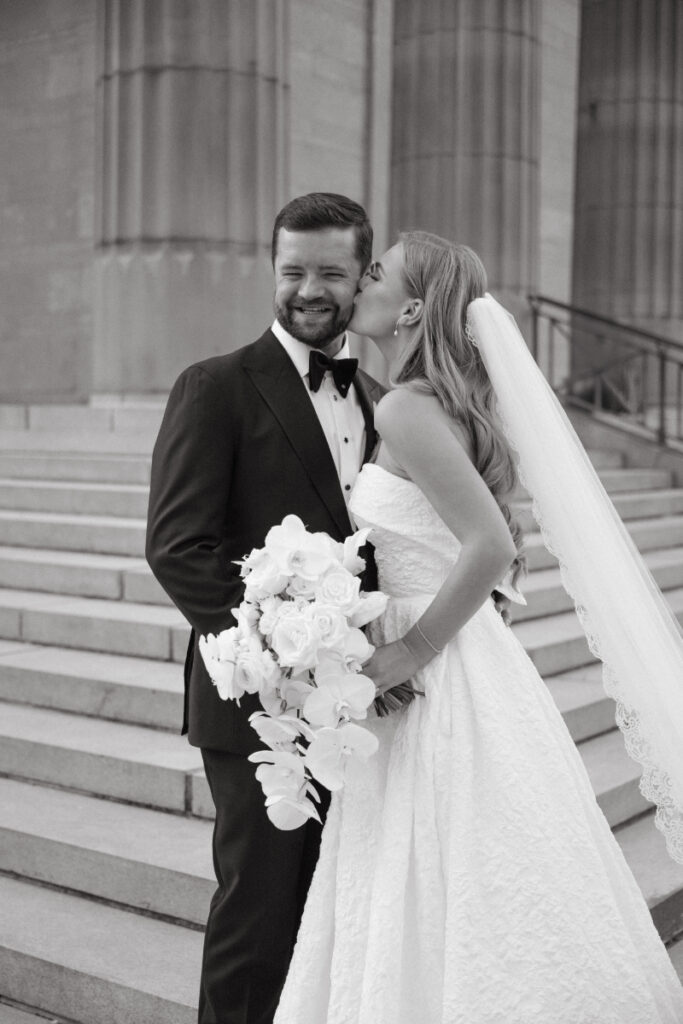 Bride kissing groom on the cheek while holding a cascading bouquet on the church steps.