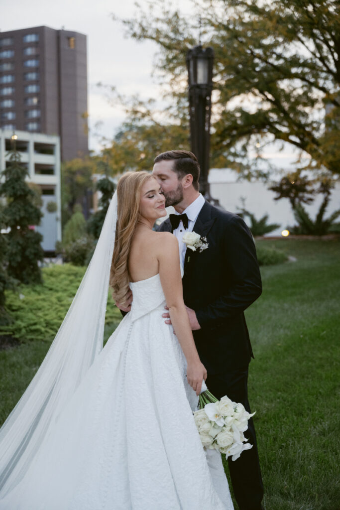 Bride and groom embracing outdoors as the groom kisses her cheek during portraits.