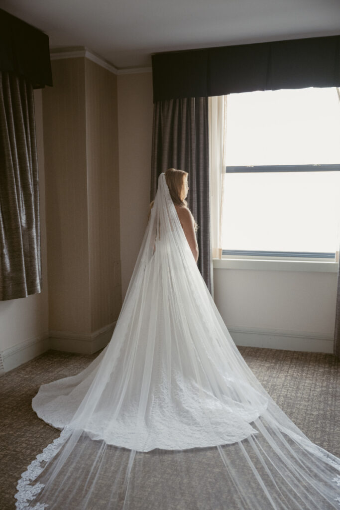 Bride standing by a window with her veil and train flowing during getting ready at a Hilton Netherland Plaza wedding.