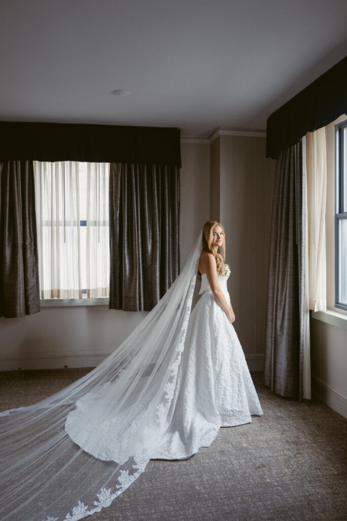 Bride standing by a window with a long flowing veil during bridal portraits at a Hilton Netherland Plaza wedding.