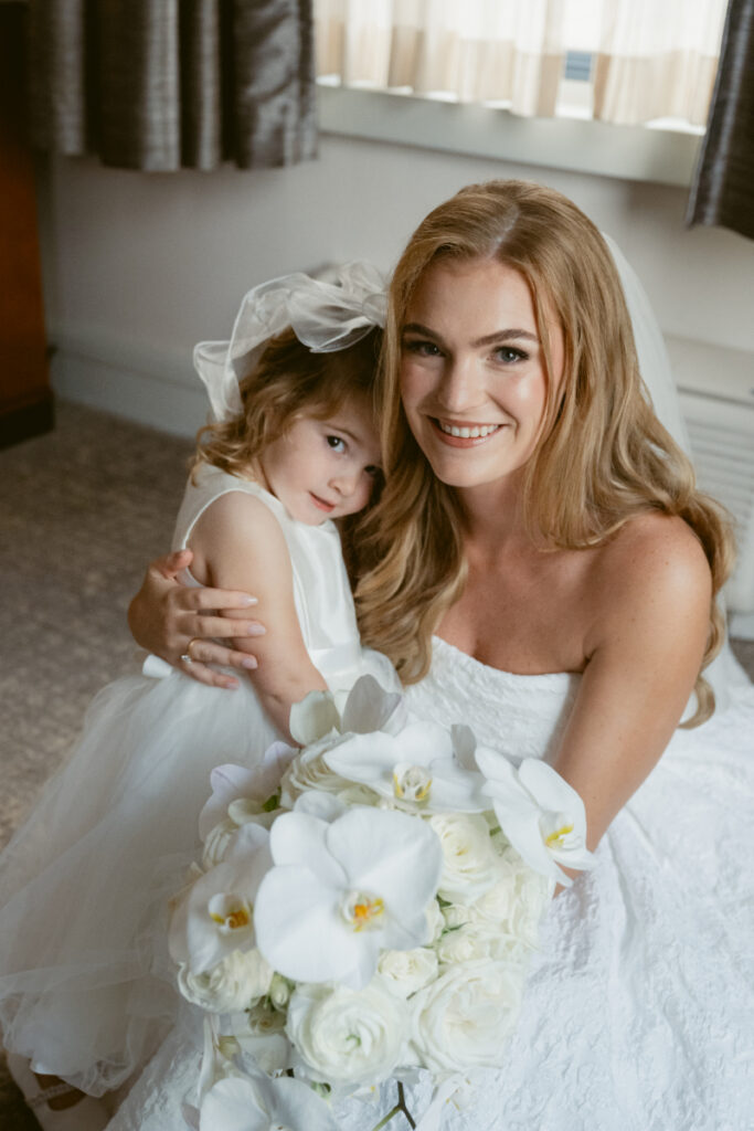 Bride smiling while holding a child and bouquet during getting ready at a Hilton Netherland Plaza wedding.