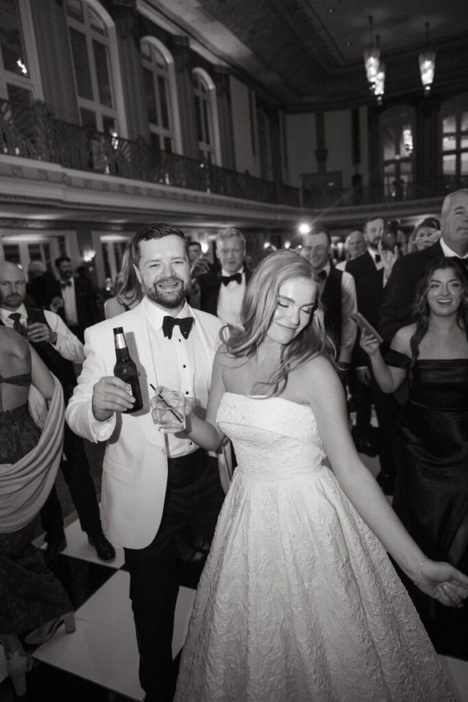 Bride and groom dancing together with drinks in hand during a lively reception moment.