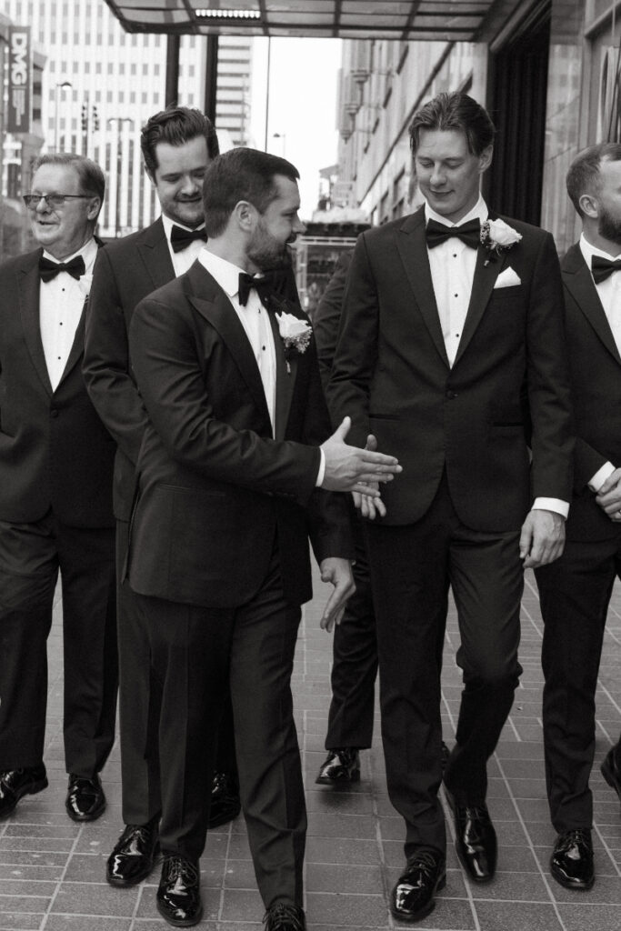 Black and white photo of groom and groomsmen in tuxedos laughing and shaking hands outdoors