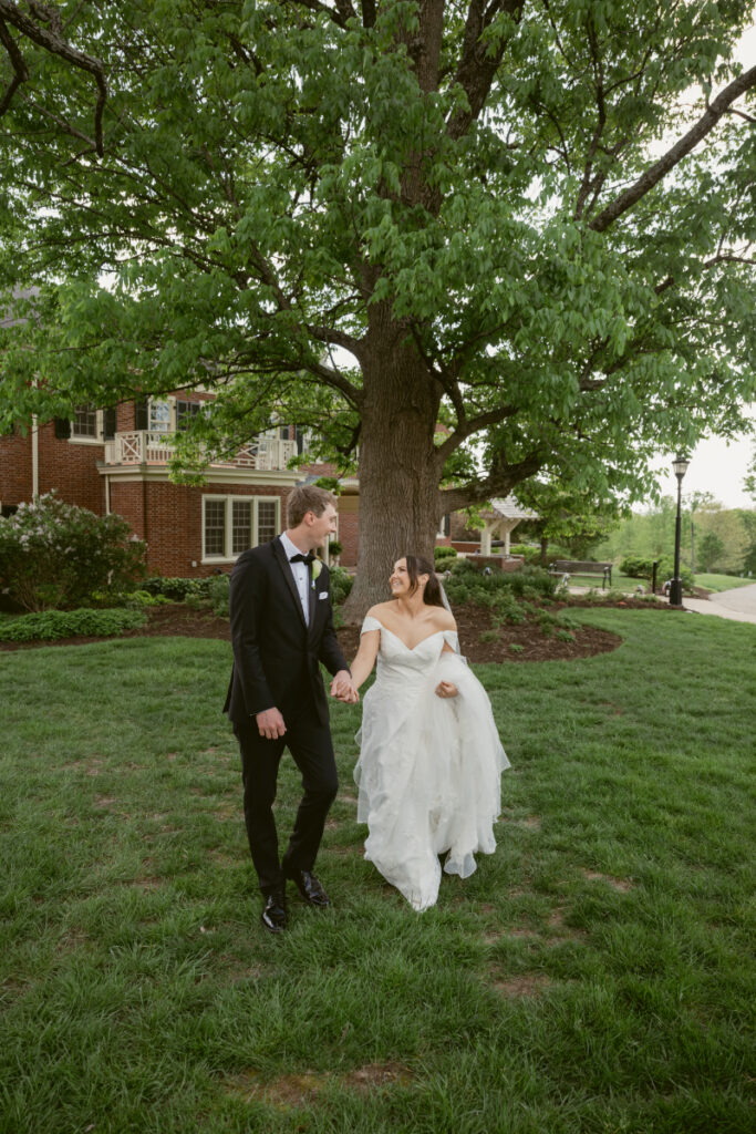 Bride and groom walk hand in hand beneath a large tree at one of Cincinnati's charming garden wedding venues.